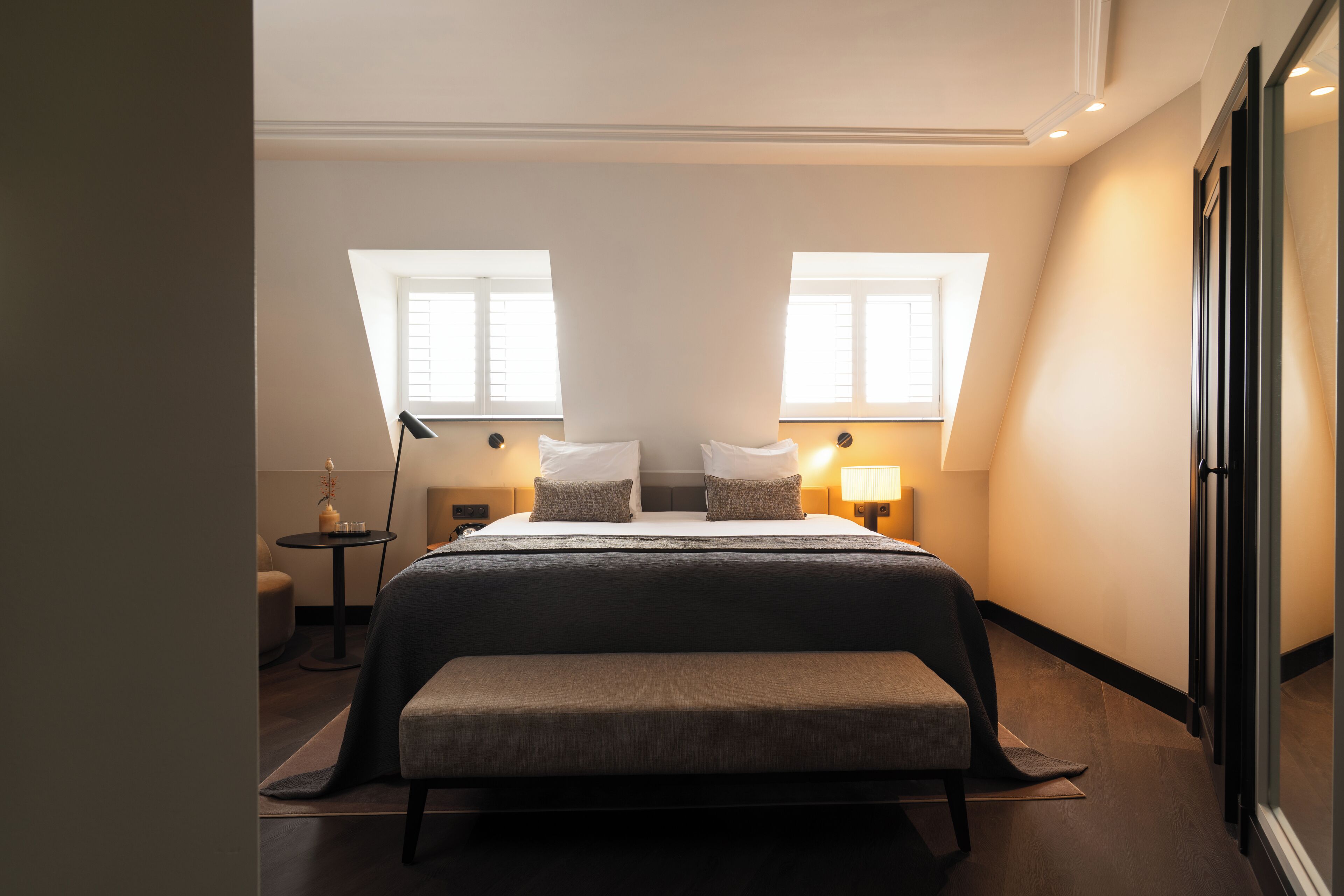 A hotel room with a large bed, two bedside tables with lamps, a bench, and two windows providing natural light.