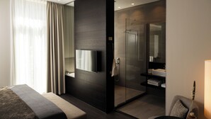 Una habitación de hotel moderna con una cama grande, televisor montado en la pared, zona de ducha y baño con lavamanos y espejo.