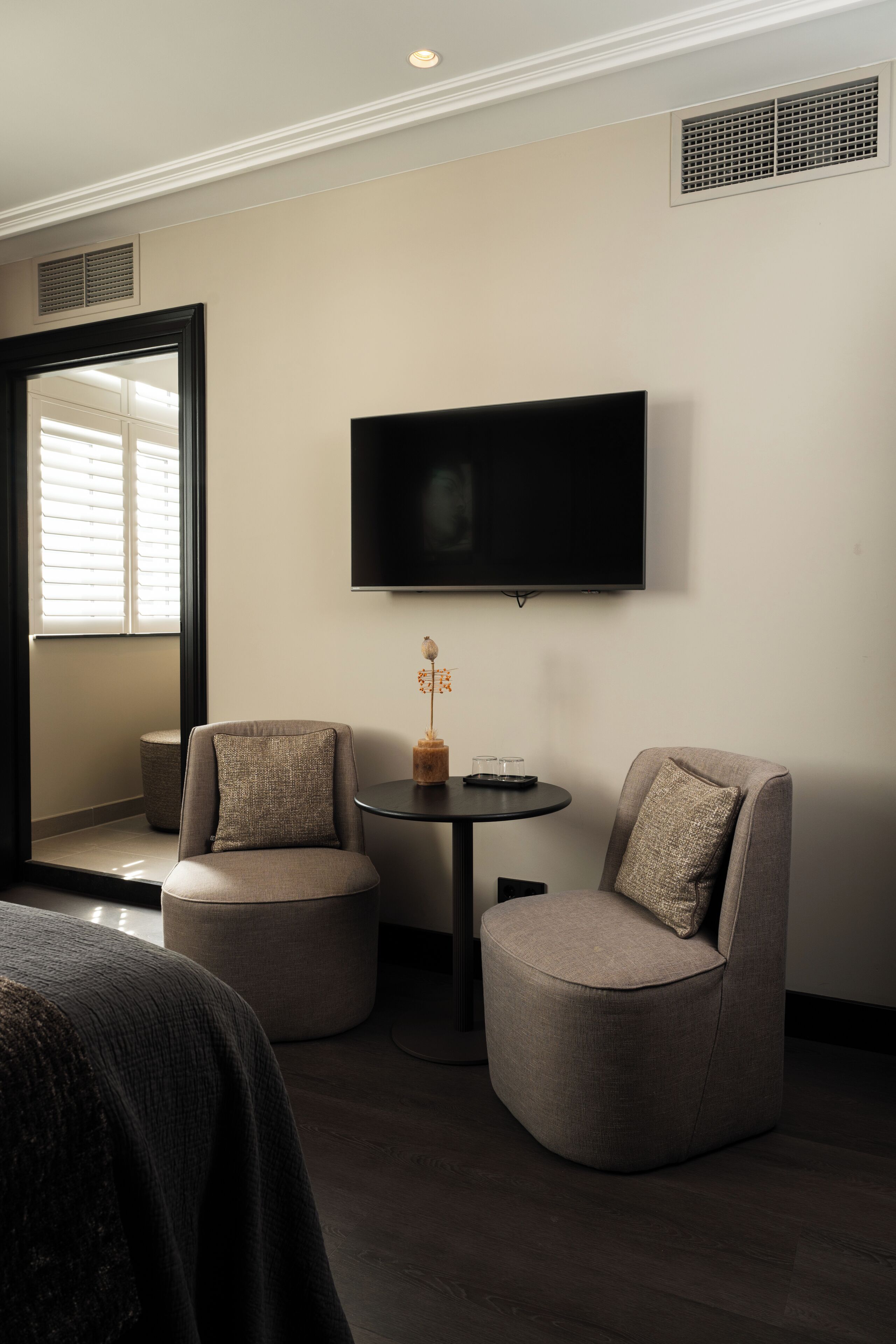 A modern hotel room with a flat-screen TV mounted on the wall, two round armchairs, a small round table with a decorative item, and a large window with blinds.
