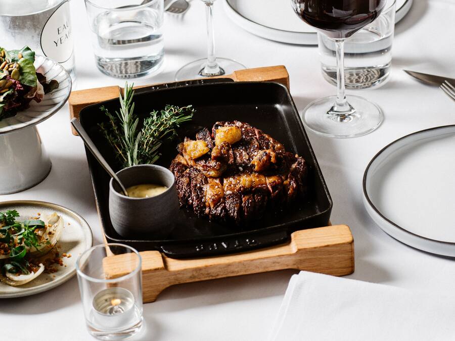 A dining table with a roasted meat dish, two glasses of red wine, and a small bowl of sauce.