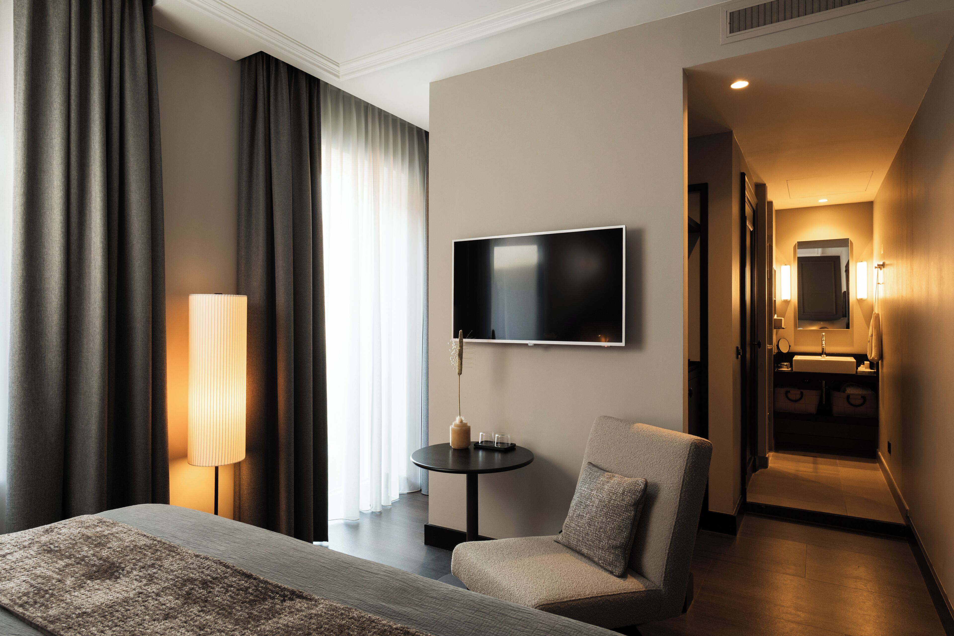 A hotel room with a bed, a chair, a TV mounted on the wall, a lamp, and a view of the hallway.