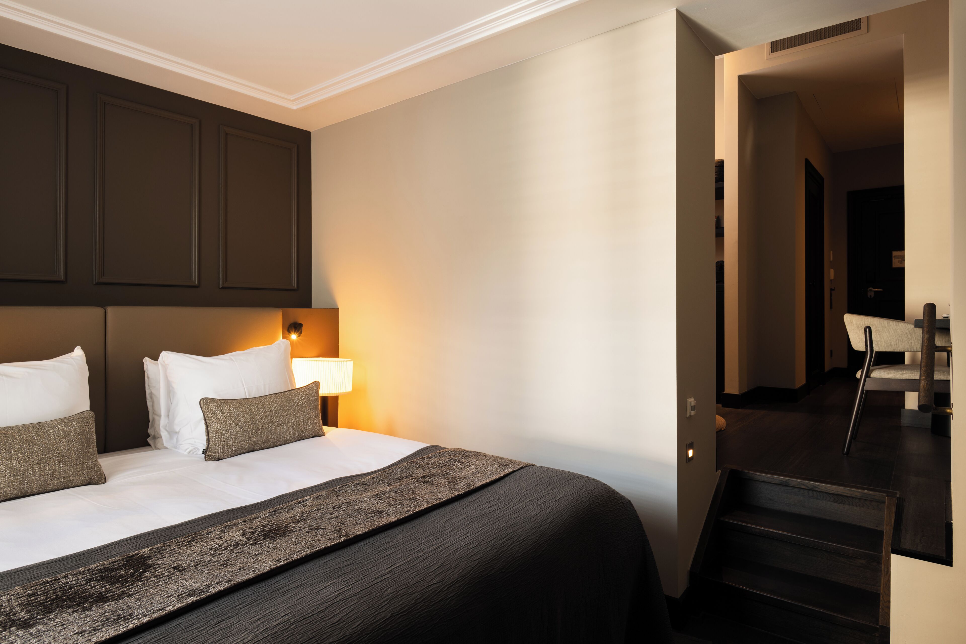 A hotel room with a large bed, a nightstand with a lamp, and a view of the hallway.