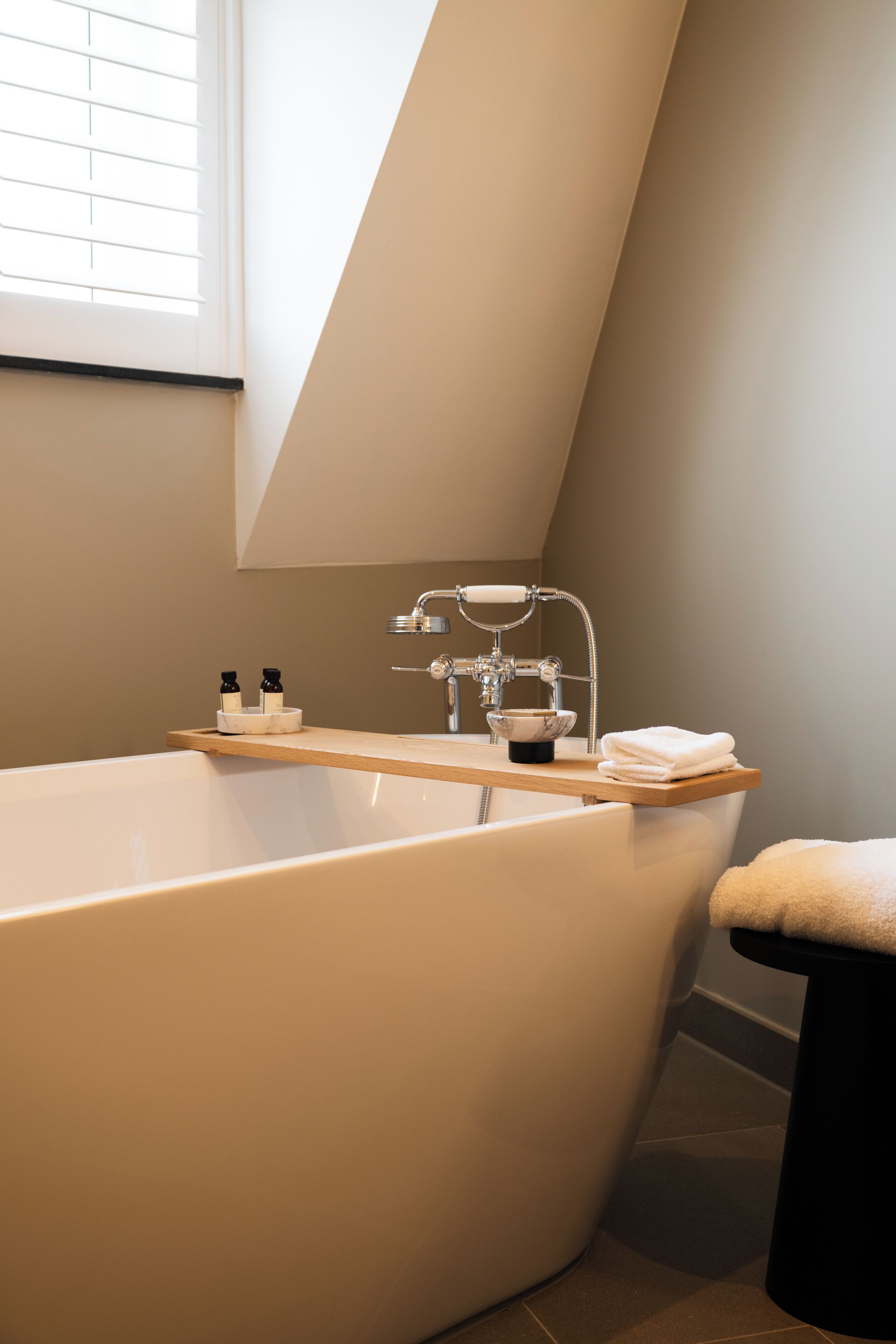 A modern bathroom with a freestanding bathtub, a wooden shelf with toiletries, and a black stool.