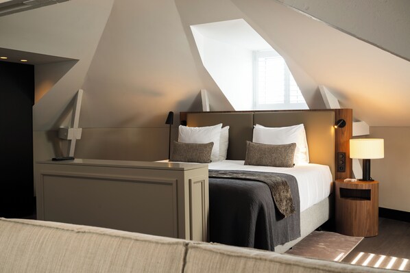 Una habitación de hotel moderna con techo inclinado, una cama grande con cabecera de madera, una mesita de noche con lámpara y vistas a través de un tragaluz.