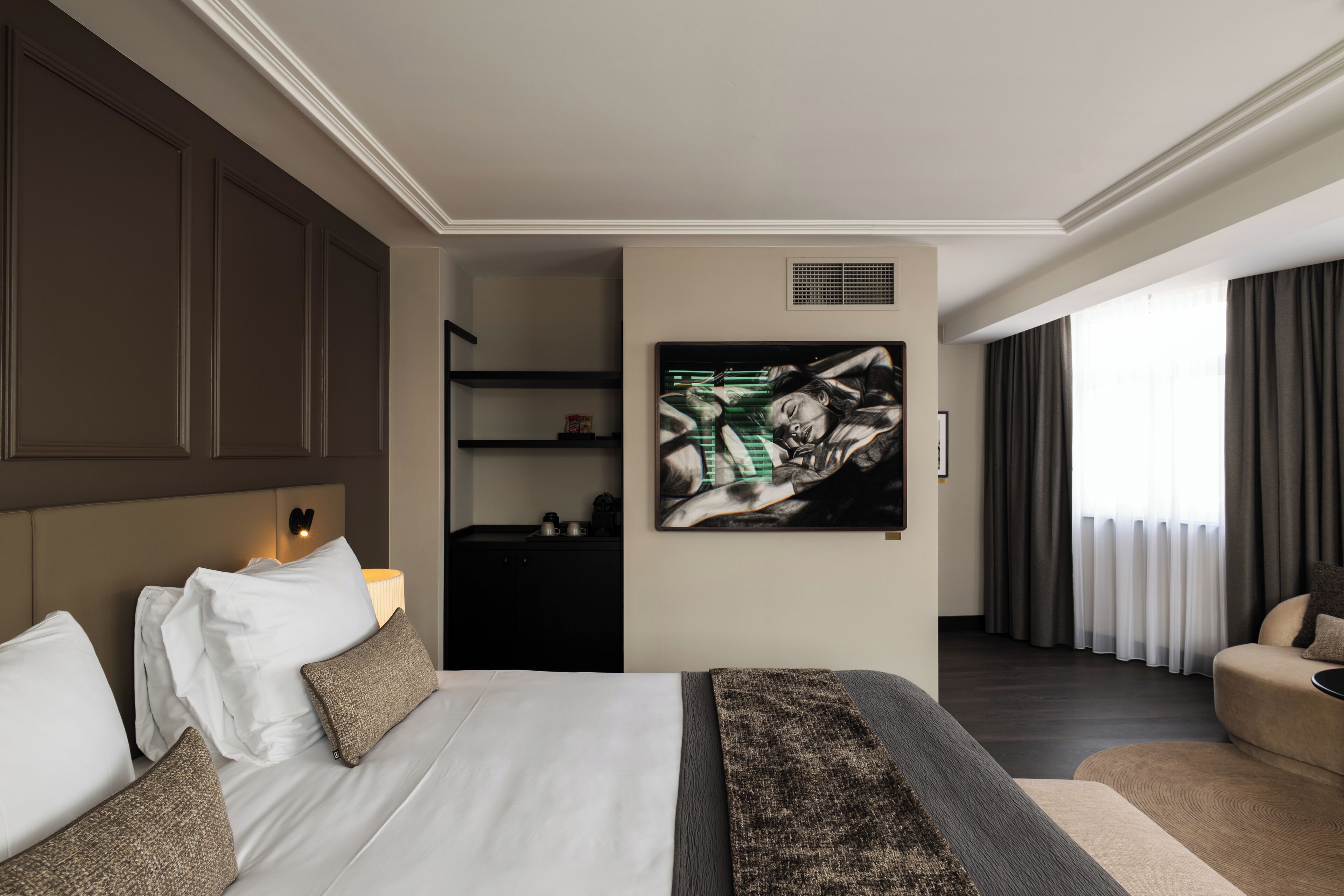 A hotel room with a large bed, a painting on the wall, a desk with a lamp, and a view of the outdoors through a window.