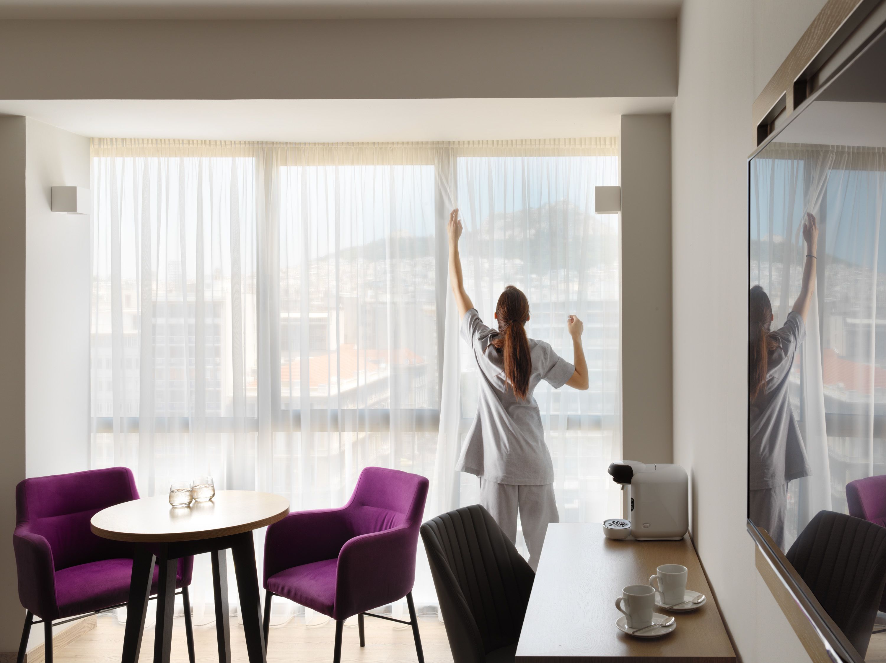A hotel room with a large window, a dining table with purple chairs, a coffee maker, and a mirror reflecting the room.