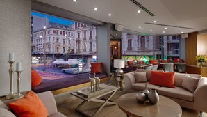 A modern hotel lobby with a large window offering a view of a city street, a seating area with sofas and a coffee table, and a bar area with a counter and seating.