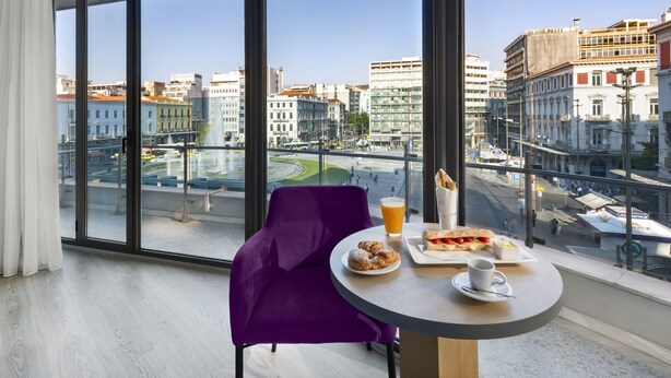 A modern room with a large window, a purple chair, a round table with breakfast items, and a city view.