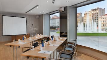 Een vergaderzaal met een lange tafel, stoelen, een scherm voor een beamer en uitzicht op een stadsbeeld met gebouwen en een fontein.