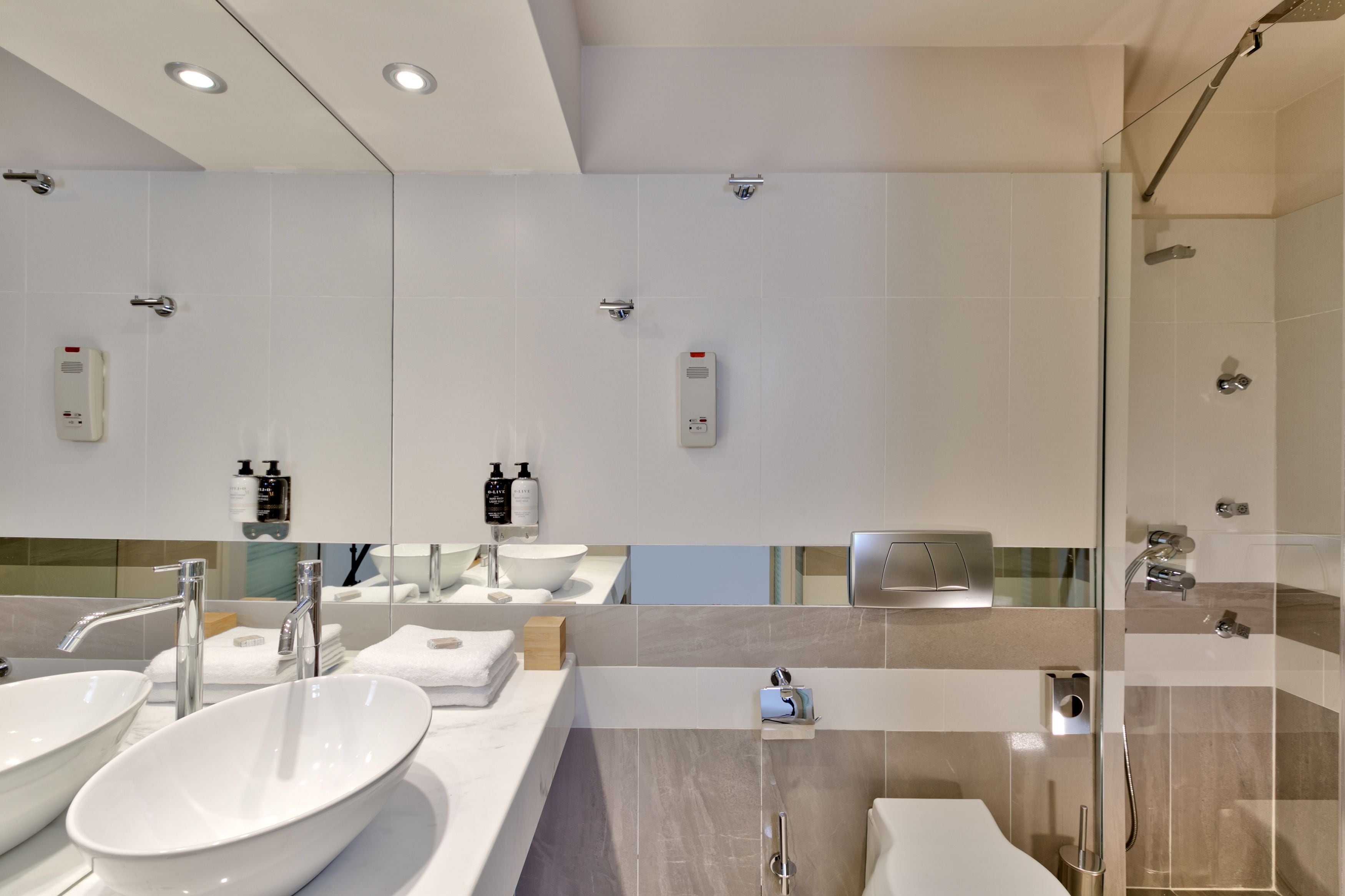 A modern bathroom with a large mirror, two sinks, a shower, and a toilet.