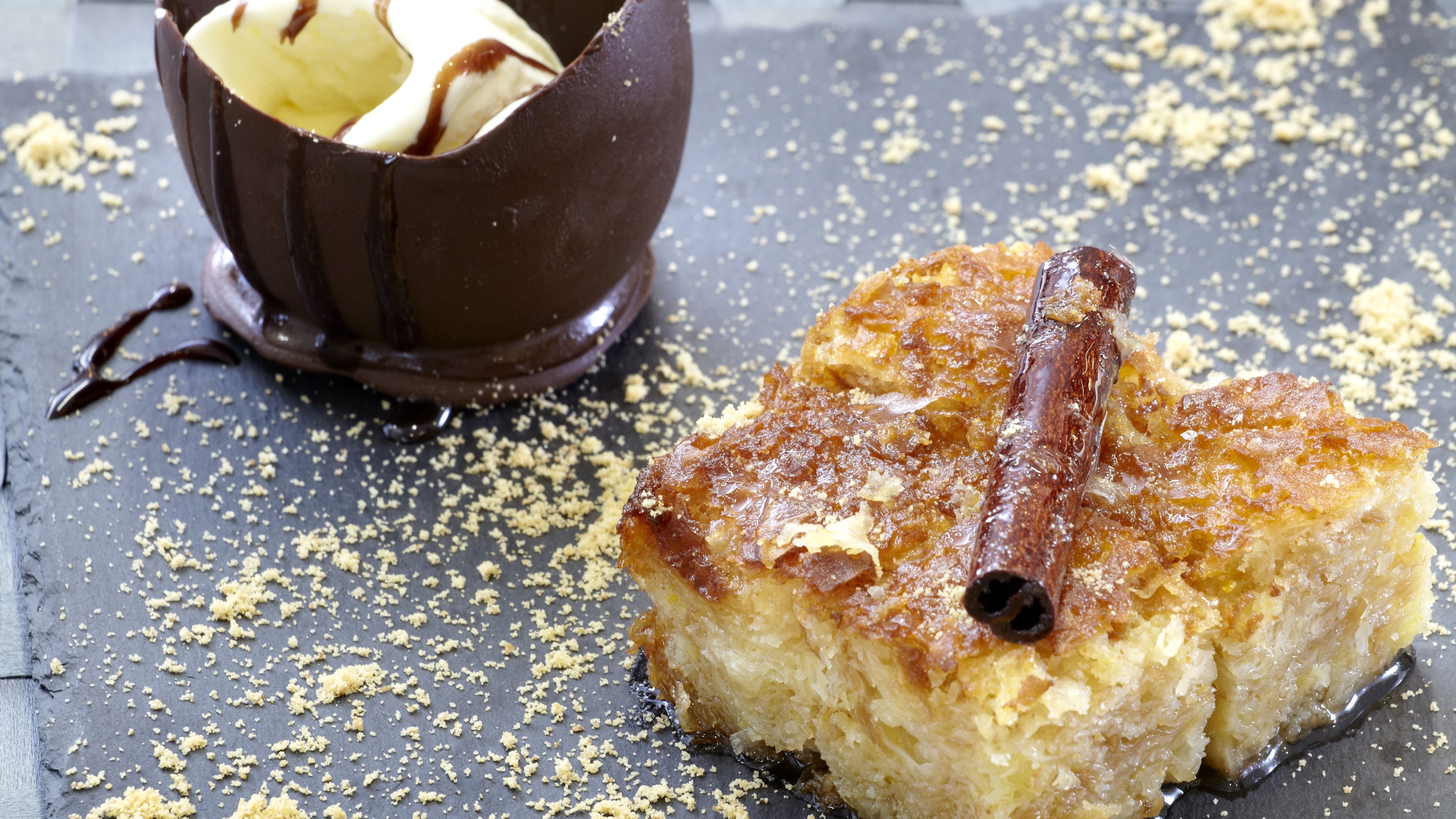 Un postre con una rama de canela encima, acompañado de una taza de chocolate y una cuchara.