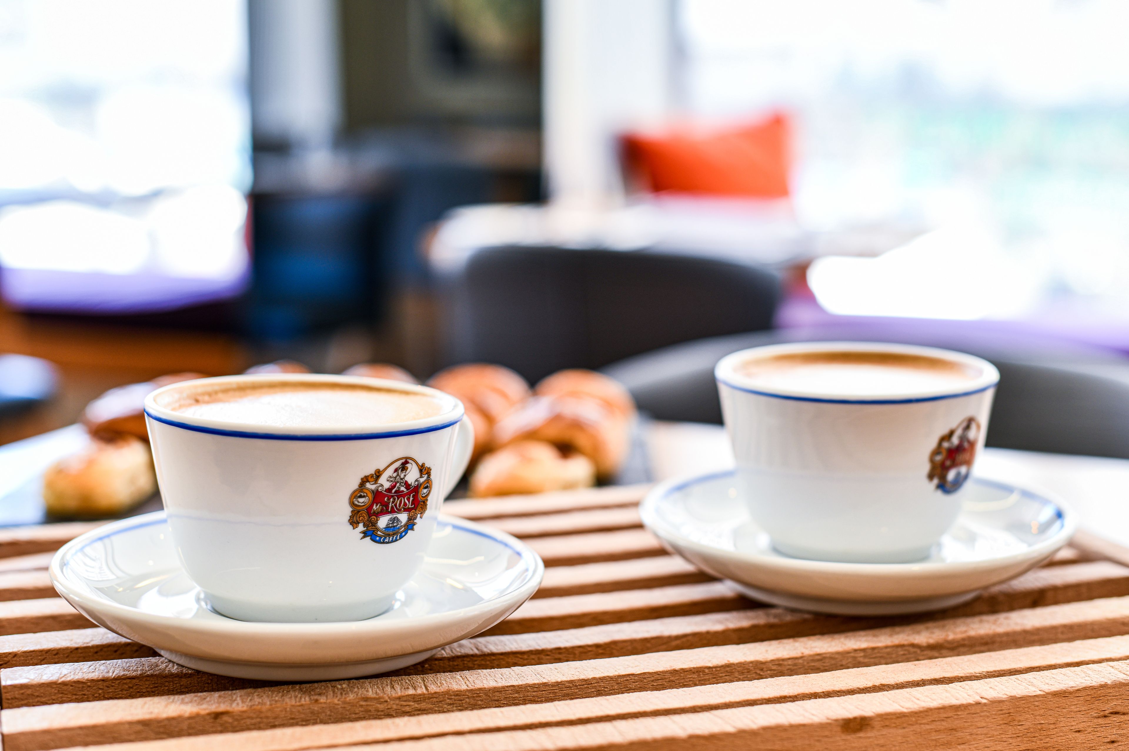 Twee kopjes koffie op een houten tafel, met gebakjes op de achtergrond.