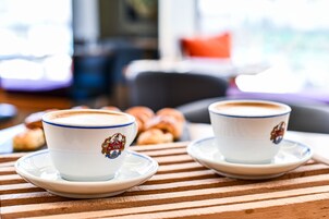 Two cups of coffee on a wooden table with pastries in the background.