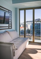 Quarto moderno com sofá, TV fixada em painel de madeira e grande porta de vidro deslizante com vista para a cidade.