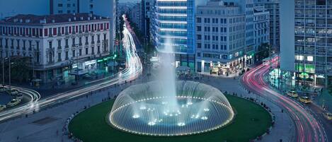 A city square with a fountain, surrounded by buildings and busy streets with light trails.