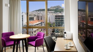 A hotel room with a large window offering a city view, including a hill with a statue. There is a round table with two purple chairs, a coffee maker, and a small table with cups and a spoon.