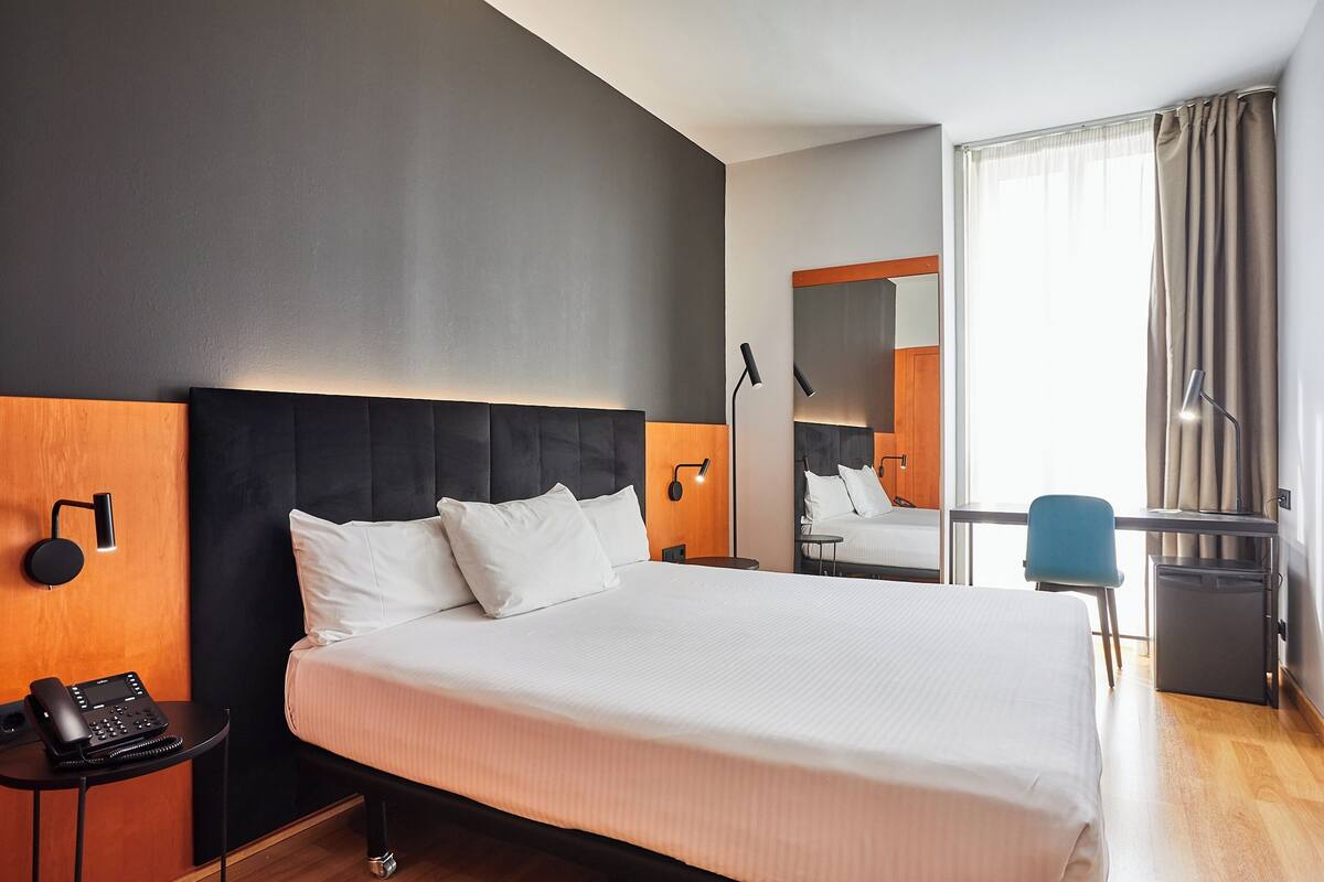 A modern hotel room with a large bed, a desk with a chair, a small refrigerator, and a telephone on a side table.