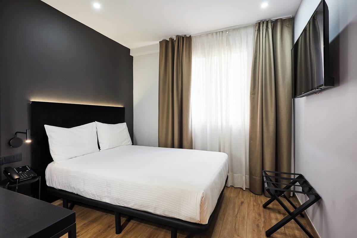 A modern hotel room with a large bed, a bedside table with a phone, a TV mounted on the wall, and a desk with a chair.