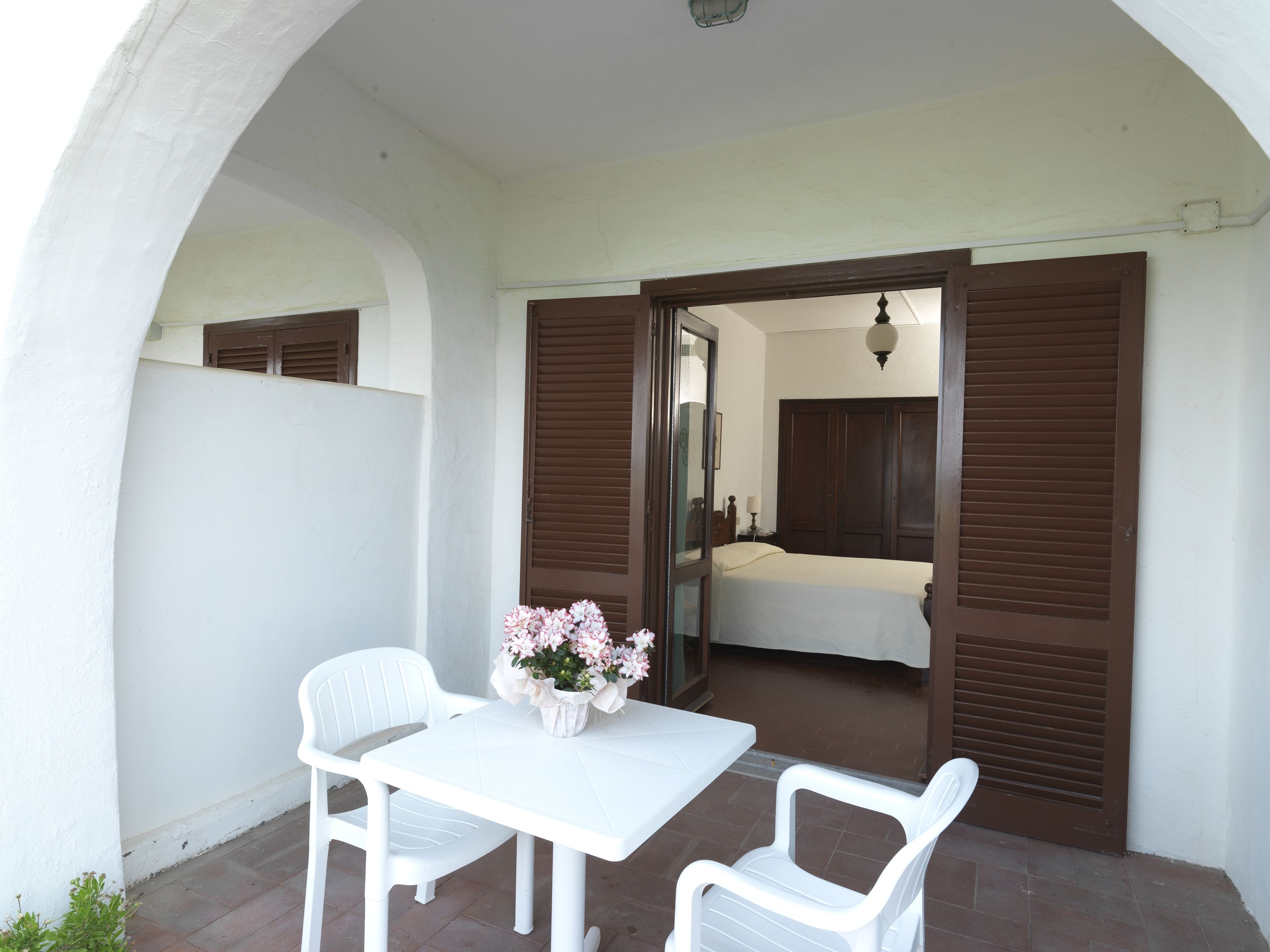 Une petite terrasse avec une table blanche et deux chaises blanches, un vase de fleurs et une vue sur une chambre à coucher avec un lit et une armoire.