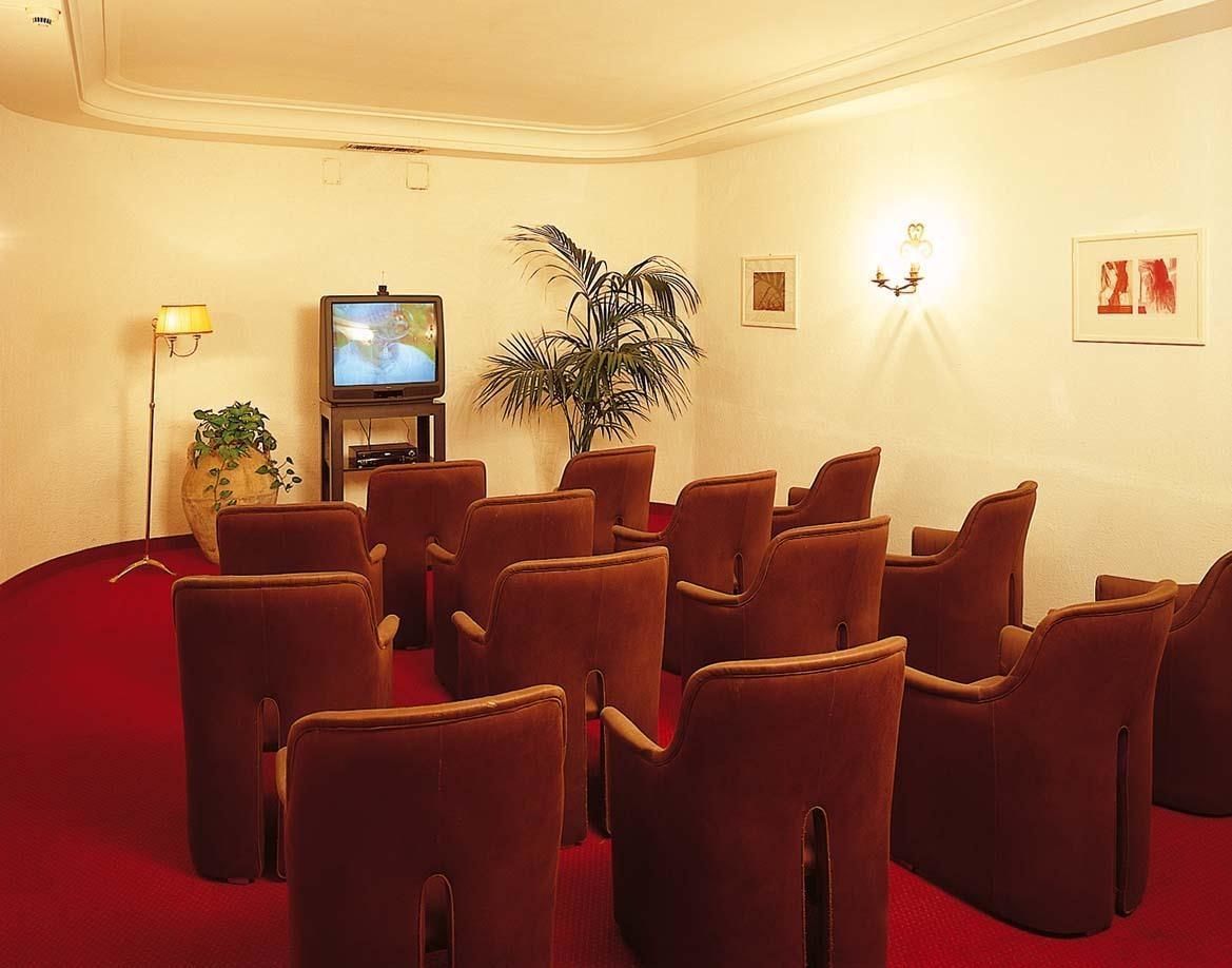 Une salle de conférence avec des rangées de chaises marron, un téléviseur posé sur un meuble, une plante en pot et des luminaires fixés au mur.
