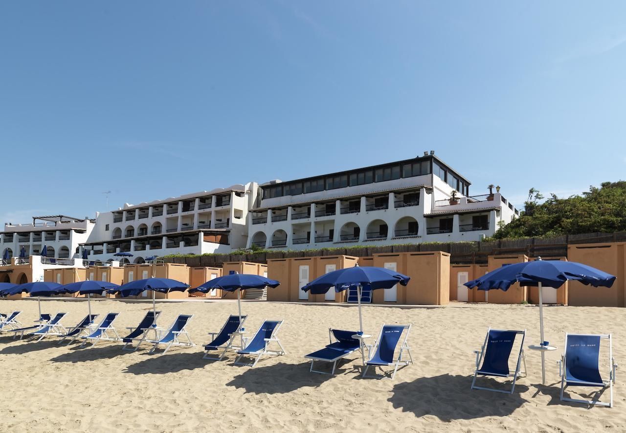 Una spiaggia con file di lettini e ombrelloni blu, e un edificio bianco con balconi sullo sfondo.