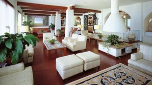 A spacious lobby with multiple seating areas, including sofas and armchairs, arranged around a central coffee table. There are large potted plants and a decorative ship model on a stand. - Hotel Le Dune (Sabaudia)