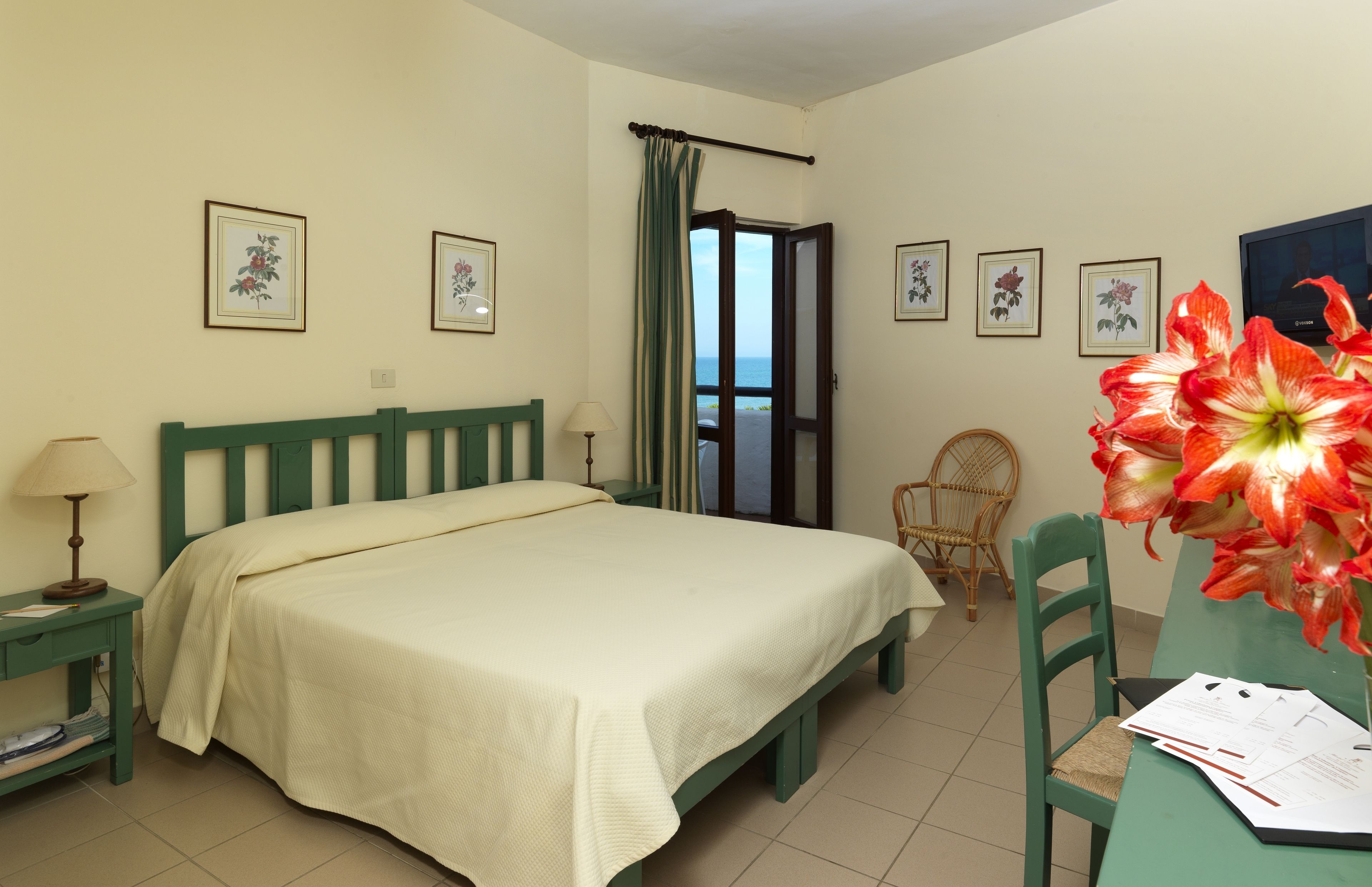 Une chambre à coucher avec un lit, une chaise, une table avec des papiers, et une vue sur la mer par la porte.