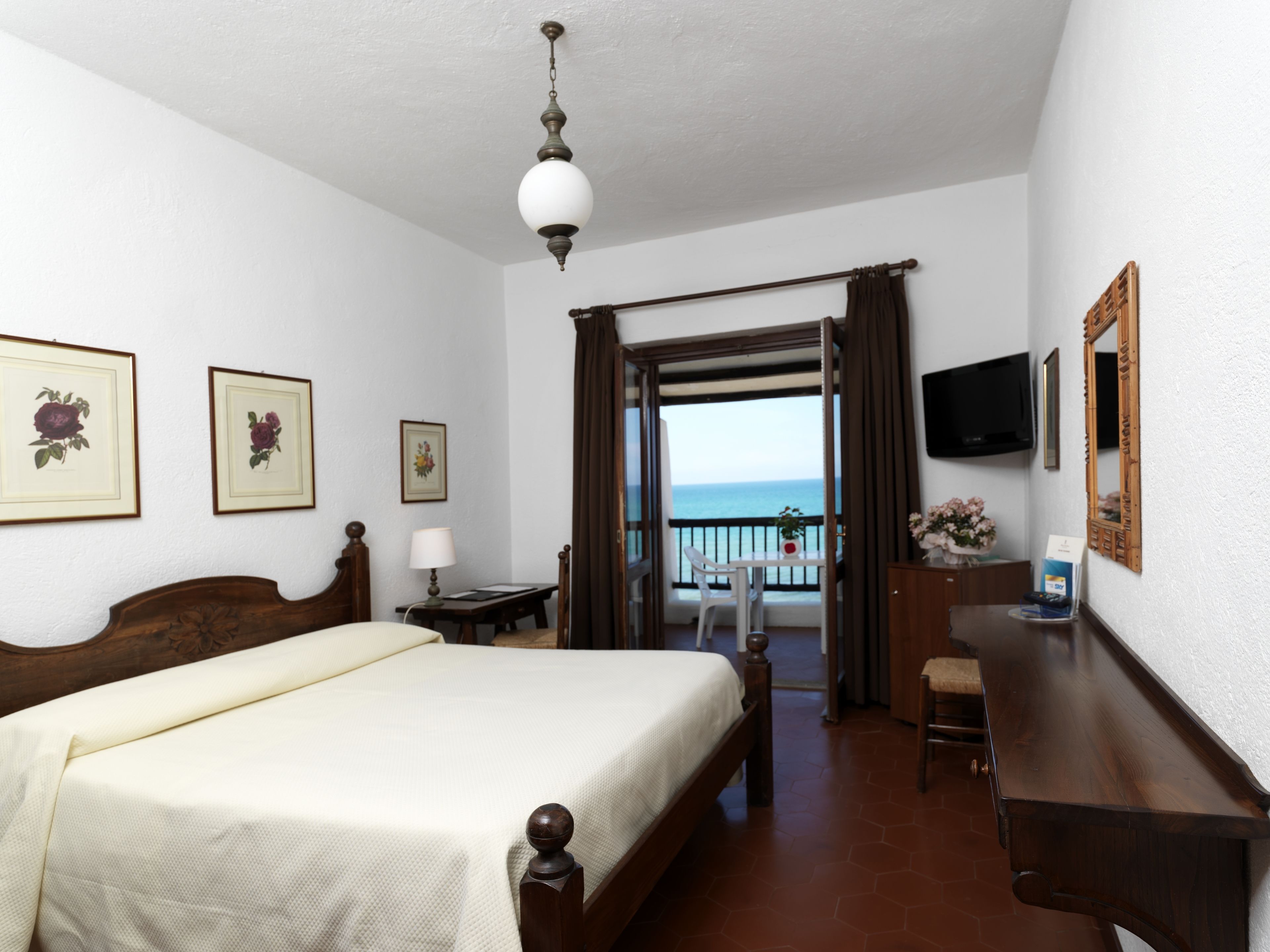 A hotel room with a bed, a desk, a chair, a TV, and a balcony with a view of the sea.