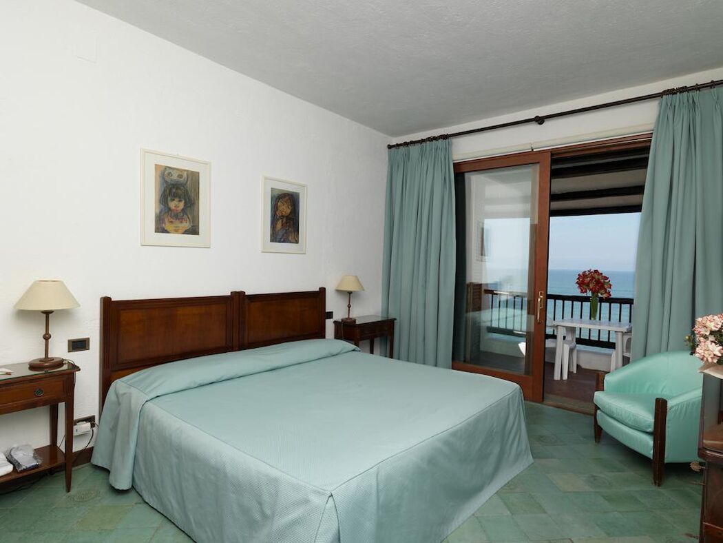 Une chambre d’hôtel avec un lit, un bureau, une chaise, un balcon donnant sur la mer et deux tableaux encadrés au mur.