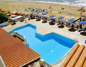 Un área de piscina con sillas de descanso y sombrillas, una bandera y una playa al fondo.