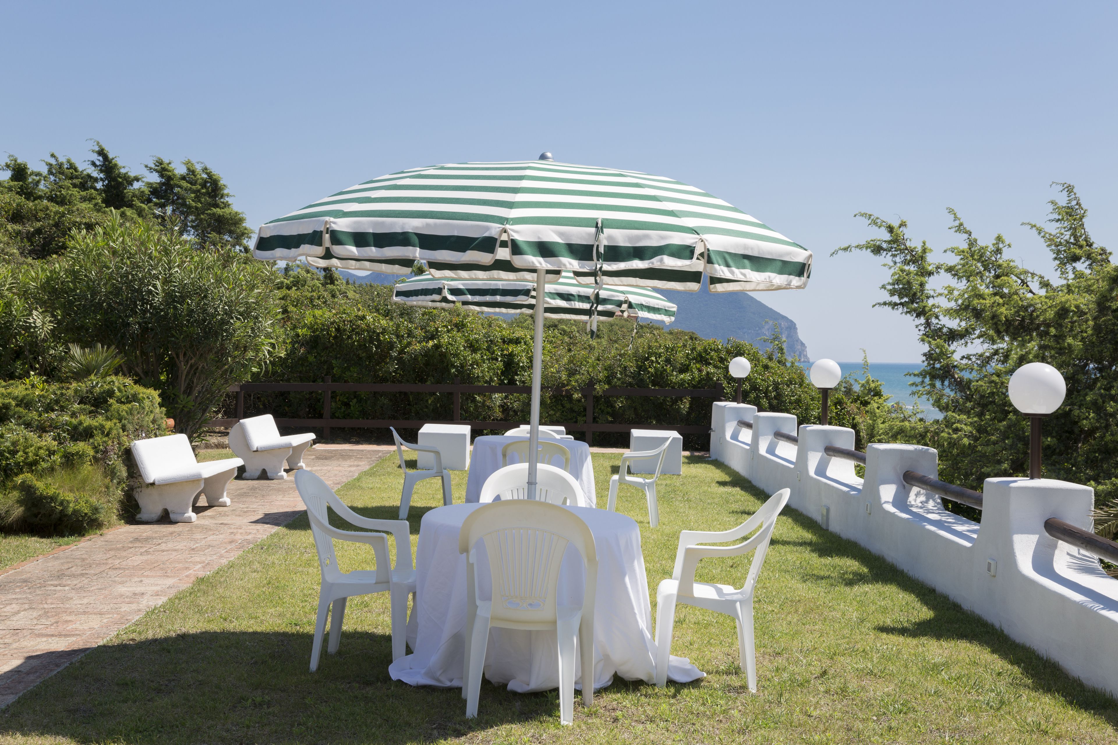 An outdoor seating area with white plastic chairs and tables, a green and white striped umbrella, and a view of the sea.