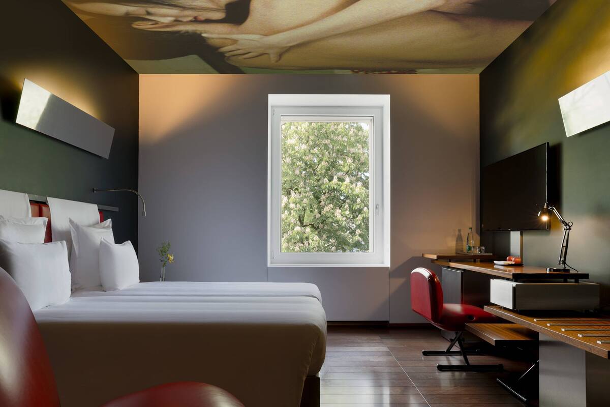 A modern hotel room with a large bed, a desk with a chair, a flat-screen TV, and a window with a view of trees.