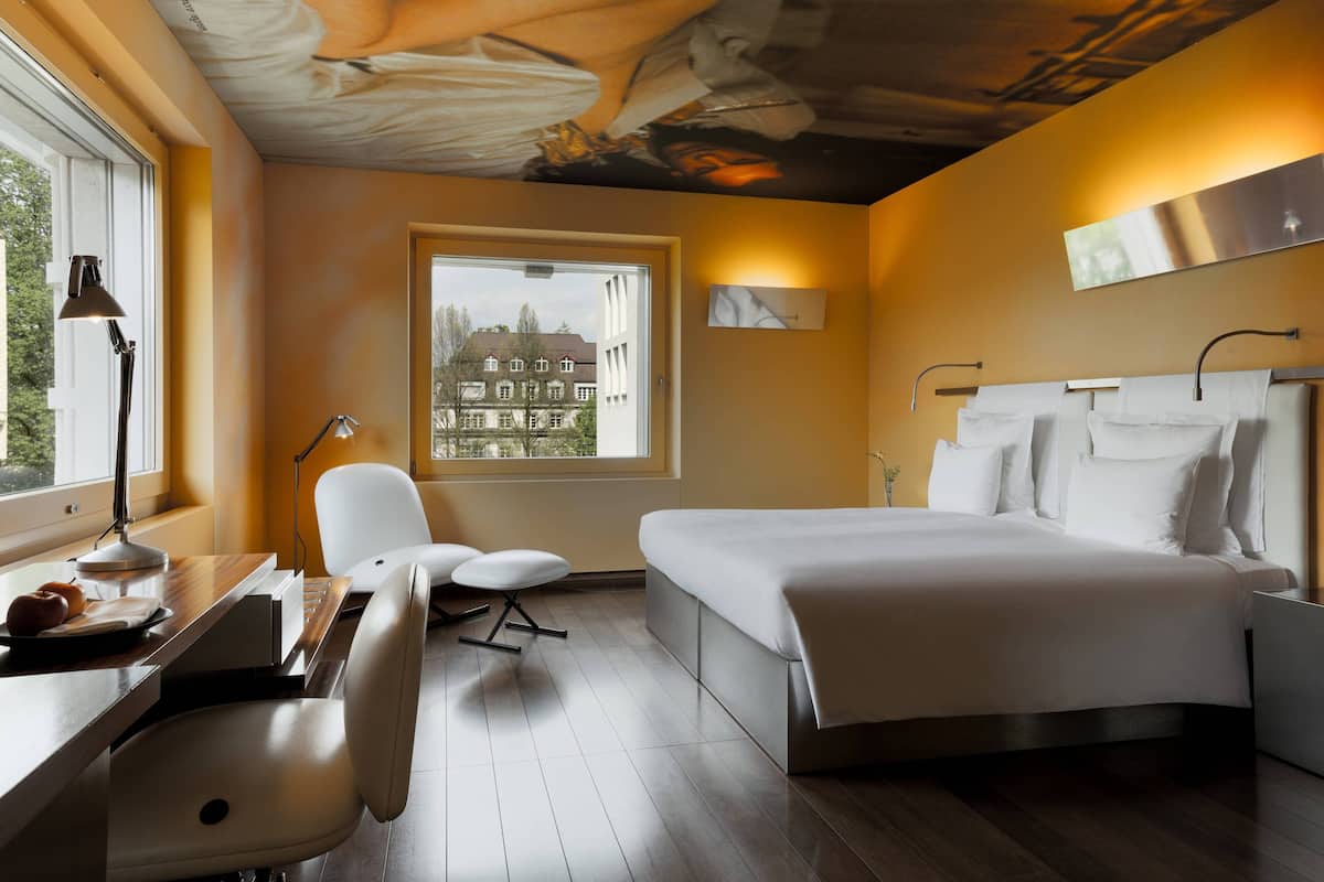 A modern hotel room with a large bed, a desk with a lamp, a chair, and a view of buildings through a window.