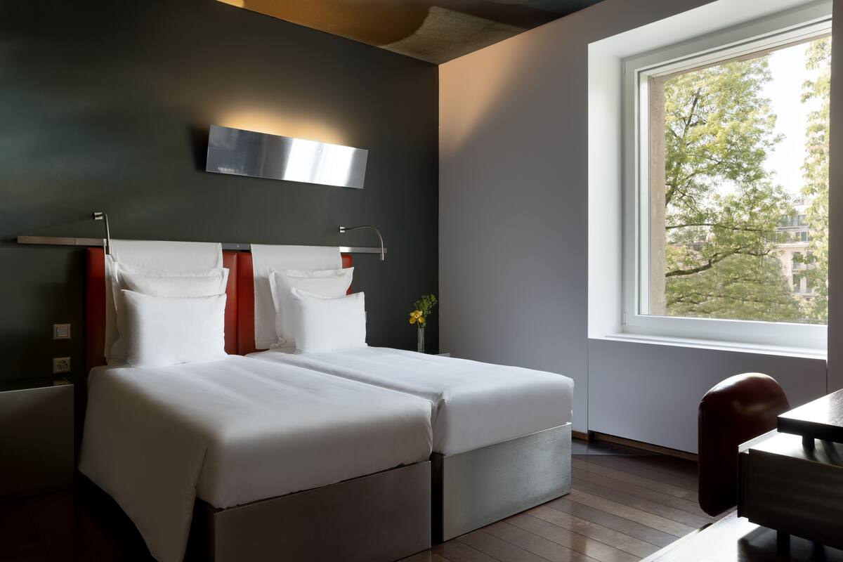 A modern hotel room with a large bed, a desk, and a window with a view of trees and buildings.