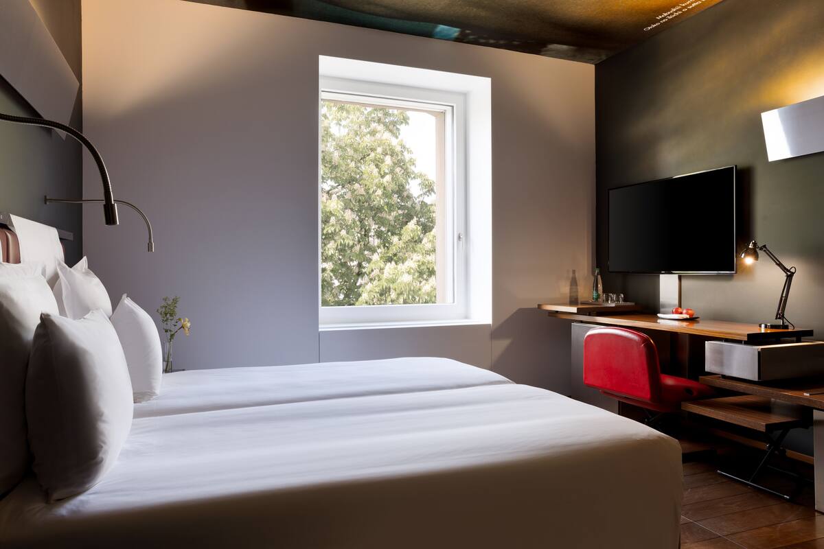 Quarto de hotel moderno com uma cama grande, uma mesa com computador, televisão e janela com vista para árvores.