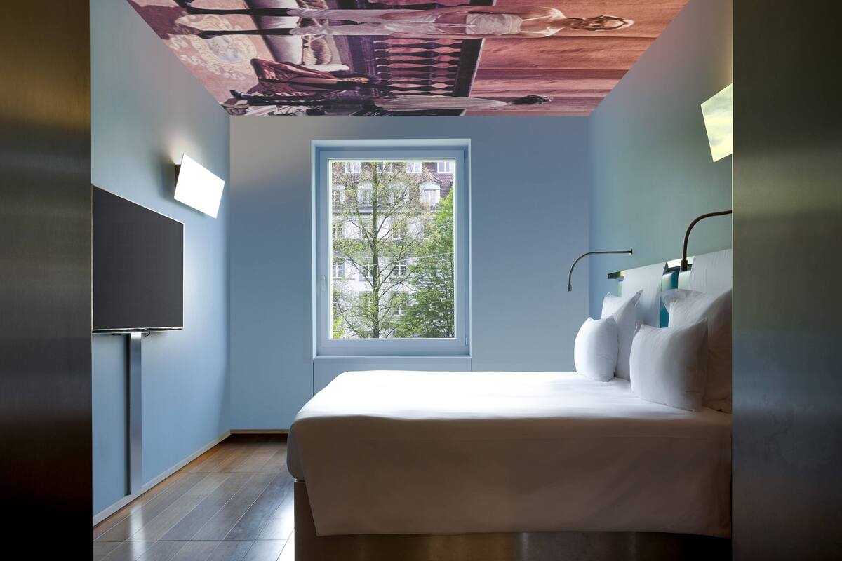 A bedroom with a bed, a window, and a flat-screen TV mounted on the wall.