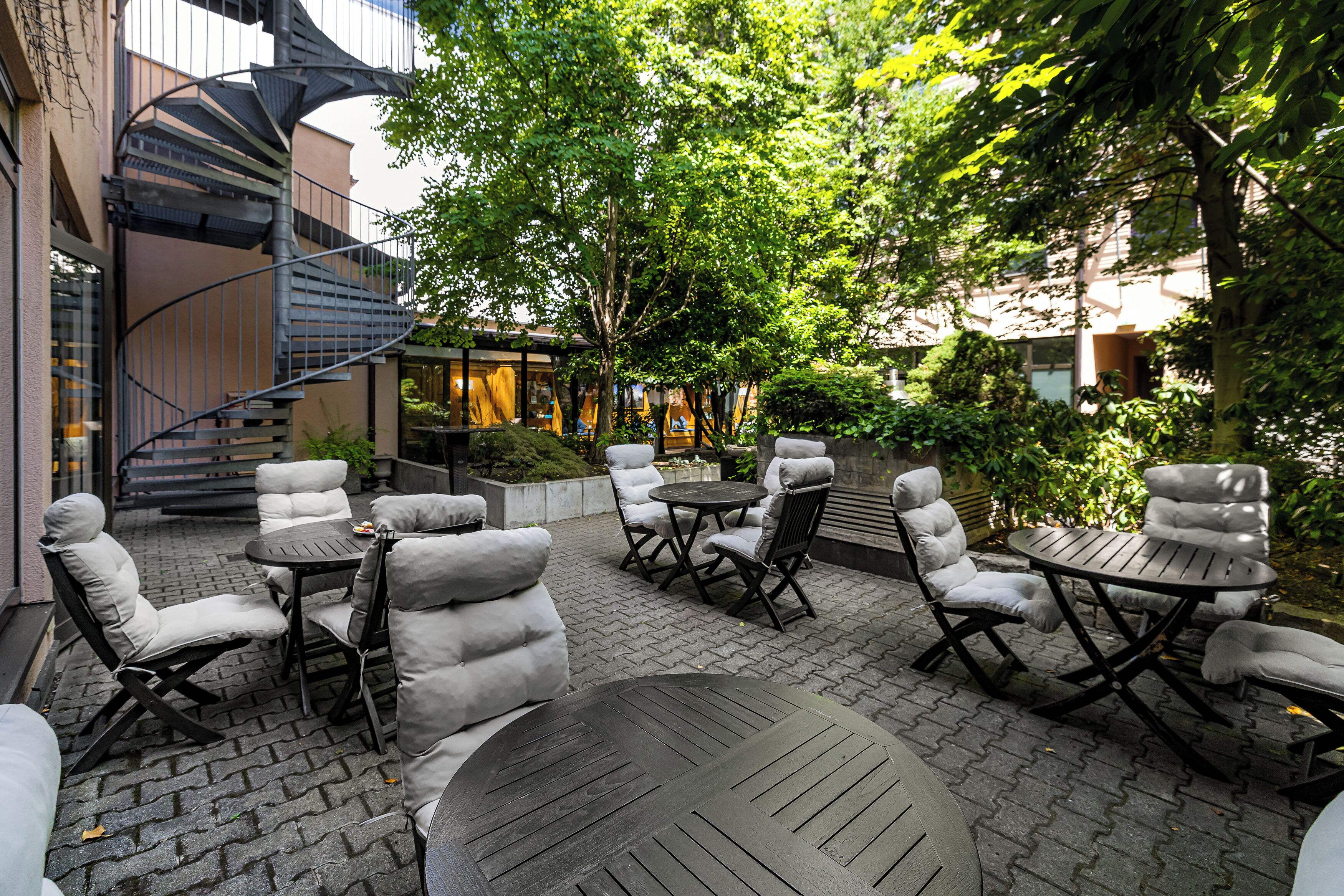 An outdoor patio with round tables and cushioned chairs, surrounded by greenery and a spiral staircase.