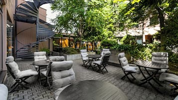 An outdoor patio with round tables and cushioned chairs, surrounded by greenery and a spiral staircase.