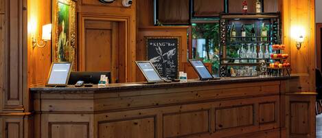 Un mostrador de recepción de madera con un reloj, un cuadro enmarco y una zona de bar con botellas y vasos.