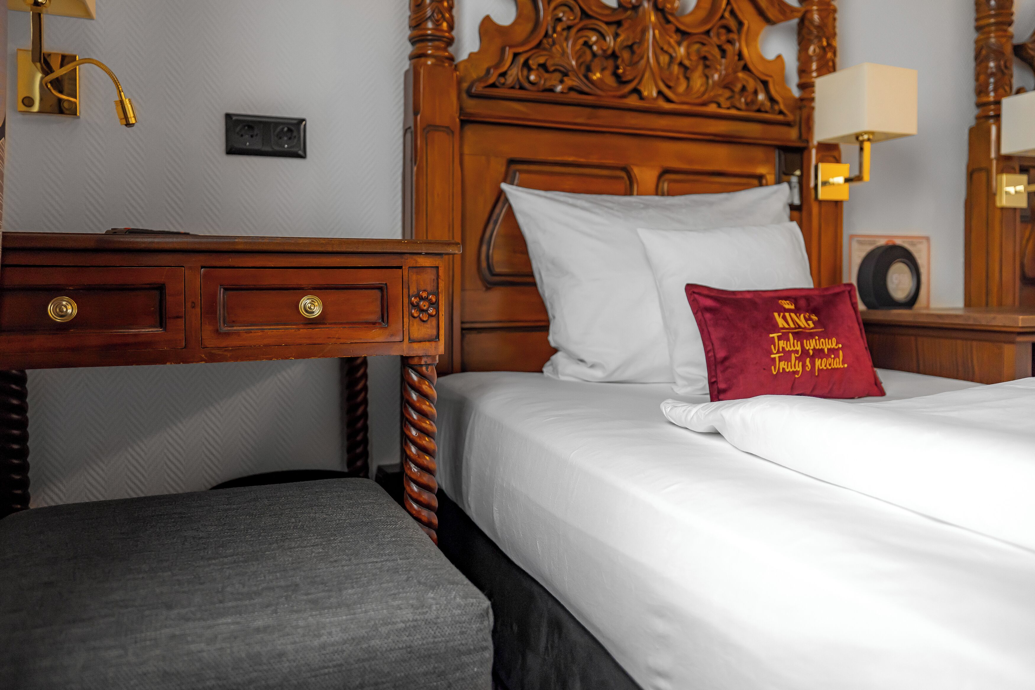 Una habitación de hotel con cabecera de madera, cama con ropa blanca, almohada roja con texto, mesita de noche y una lámpara.