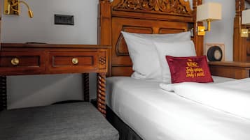 Una habitación de hotel con cabecera de madera, cama con ropa blanca, almohada roja con texto, mesita de noche y una lámpara.