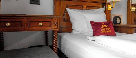 Una habitación de hotel con cabecera de madera, cama con ropa blanca, almohada roja con texto, mesita de noche y una lámpara.