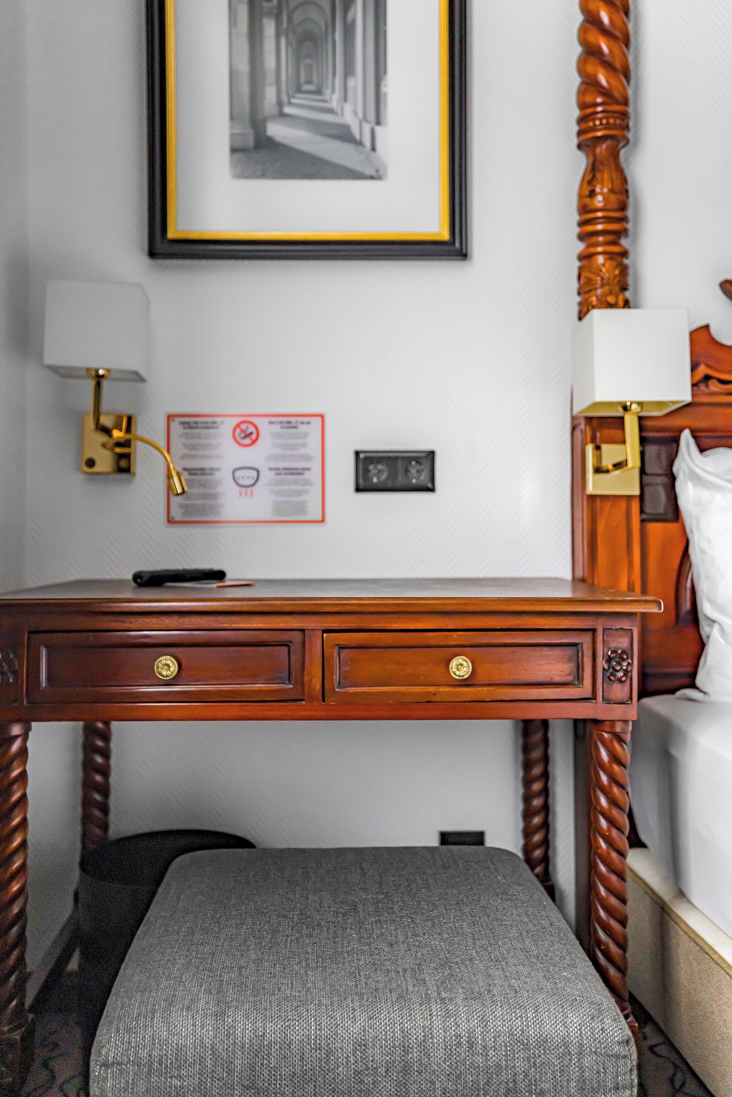 Quarto de hotel com escrivaninha de madeira, quadro decorativo na parede, mesa de cabeceira e cama com cabeceira de madeira.