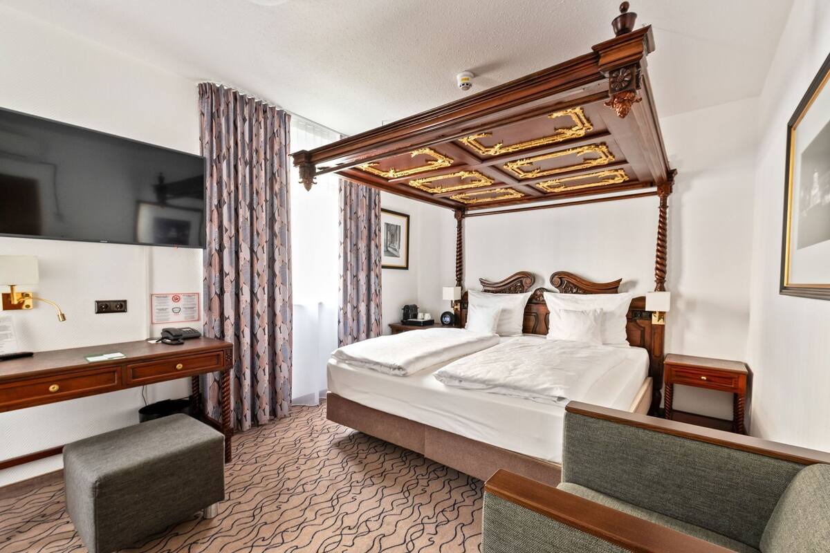 Habitación de hotel con cama con dosel, un escritorio con silla, un televisor y una alfombra estampada.