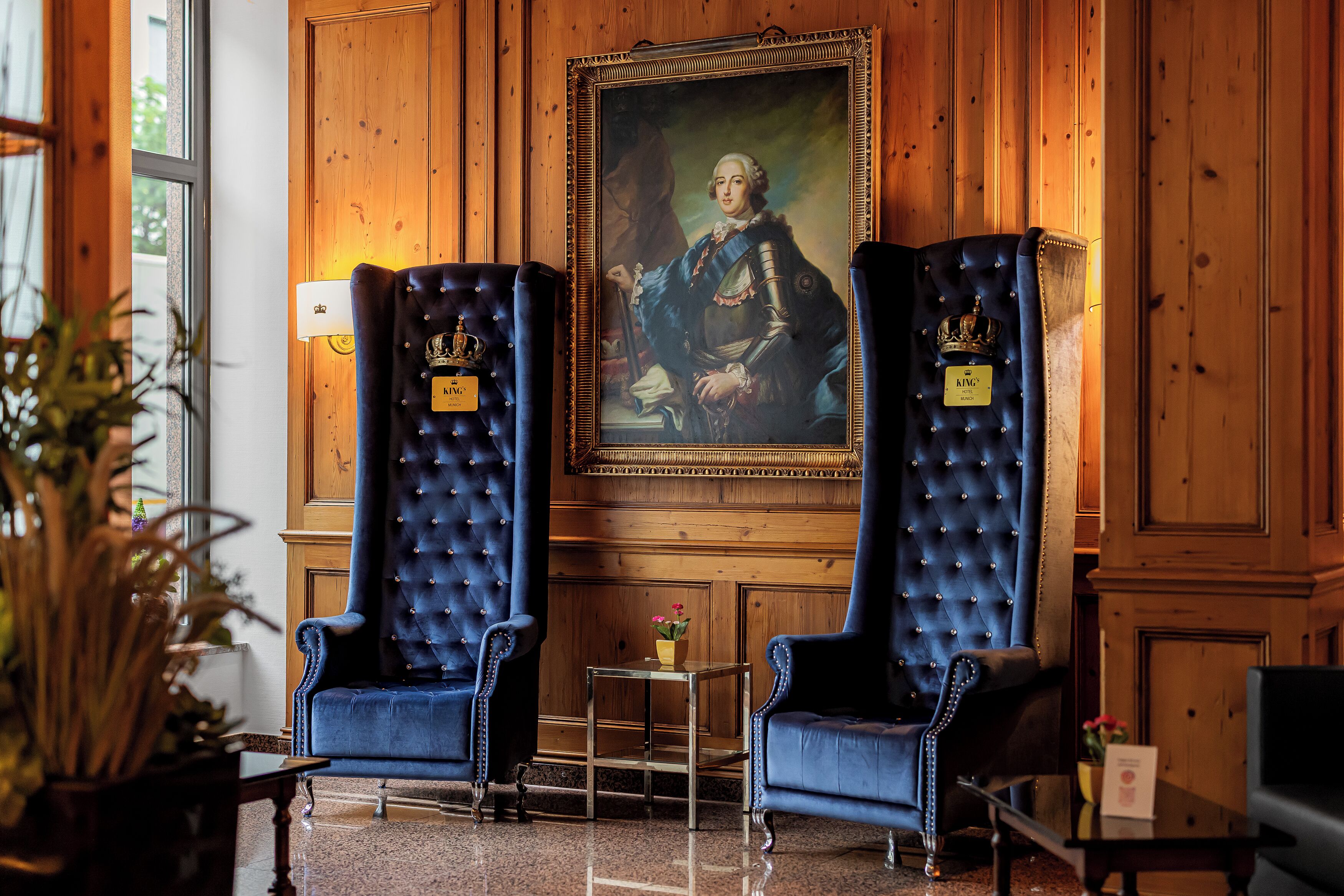 A room with two large, dark blue upholstered chairs with crown-shaped plaques, a framed portrait on the wall, and a small table with a vase of flowers.