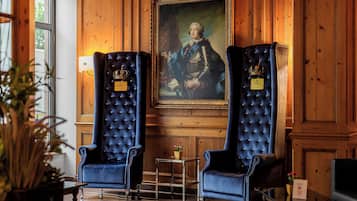 A room with two large, dark blue upholstered chairs with crown-shaped plaques, a framed portrait on the wall, and a small table with a vase of flowers.