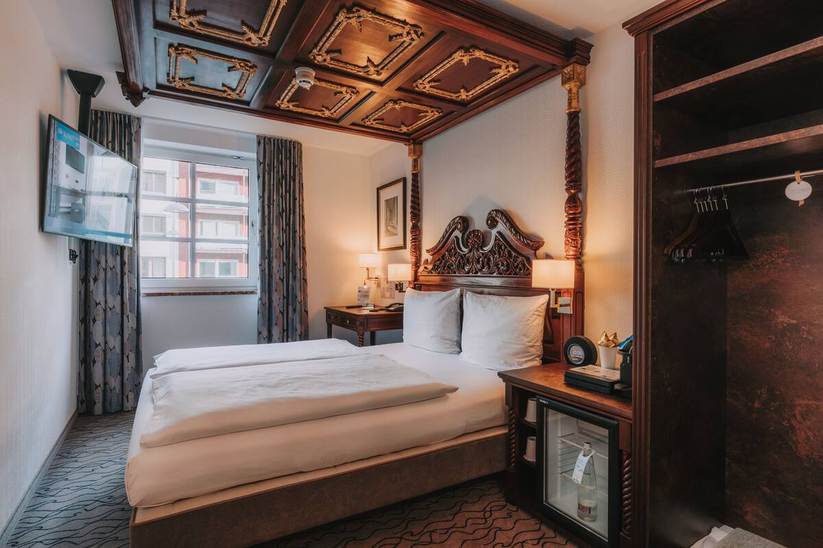 Habitación de hotel con una cama grande, una mesita de noche con lámpara, un armario y un televisor en la pared.