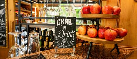 Zona de bar con un letrero que dice 'GRAB A Drink', una presentación de frutas con manzanas y una estantería de vinos al fondo.