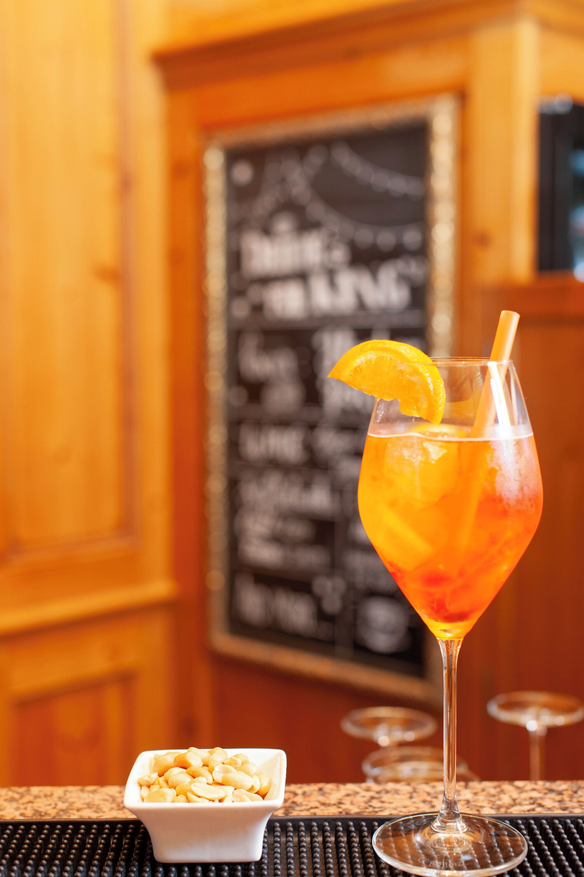 Una copa de cóctel con una rodaja de naranja, un cuenco pequeño de frutos secos y un panel de menú al fondo.