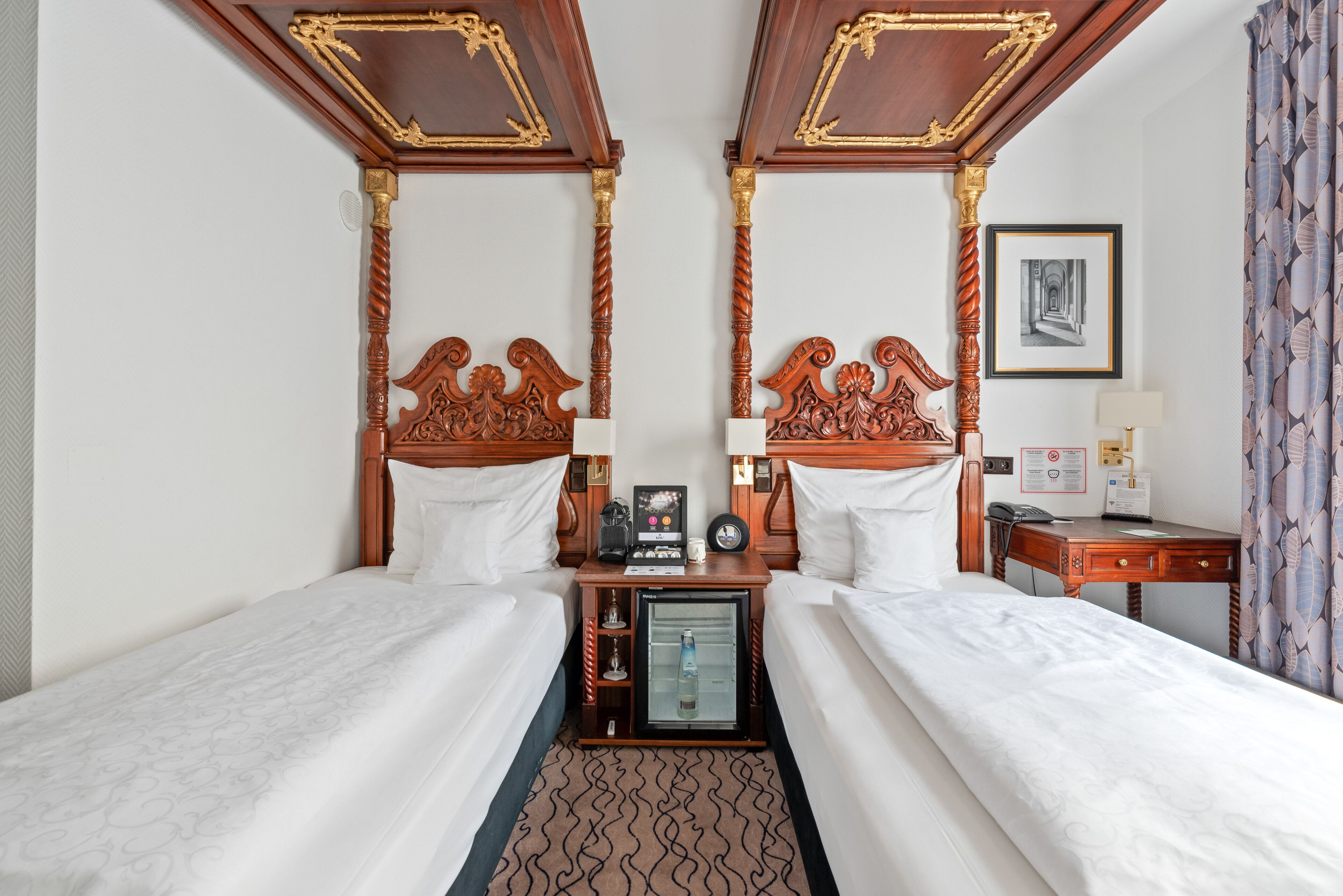 A hotel room with two beds, a minibar, a desk, and a framed picture on the wall.